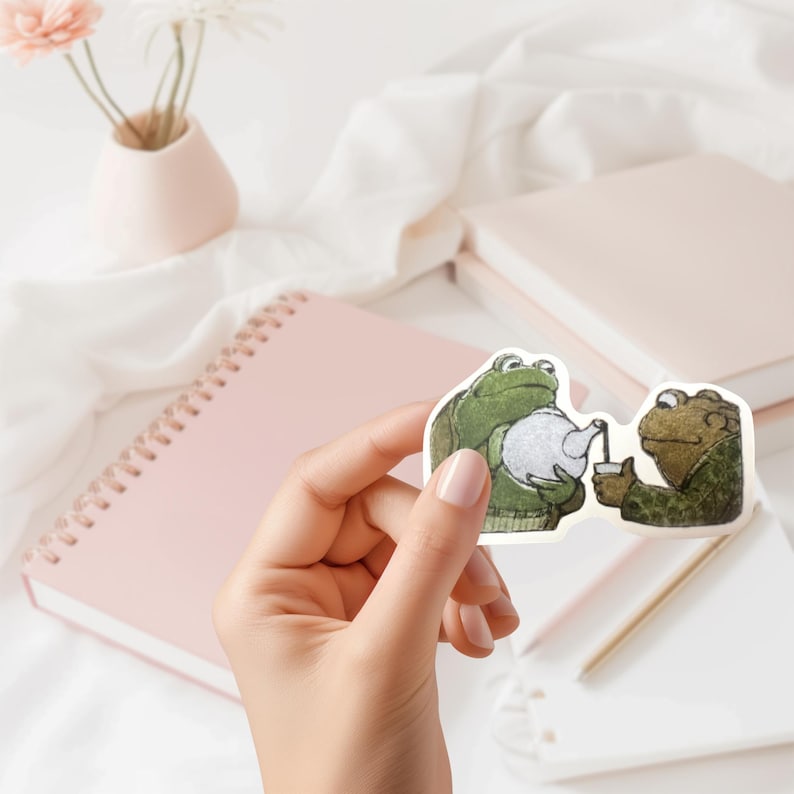 Puede incluir: Una pegatina con dos ranas de dibujos animados. Una rana vierte t&eacute; de una tetera blanca en una taza que sostiene la otra. Las ranas llevan jers&eacute;is verdes. En el fondo hay un cuaderno rosa y un jarr&oacute;n con flores.