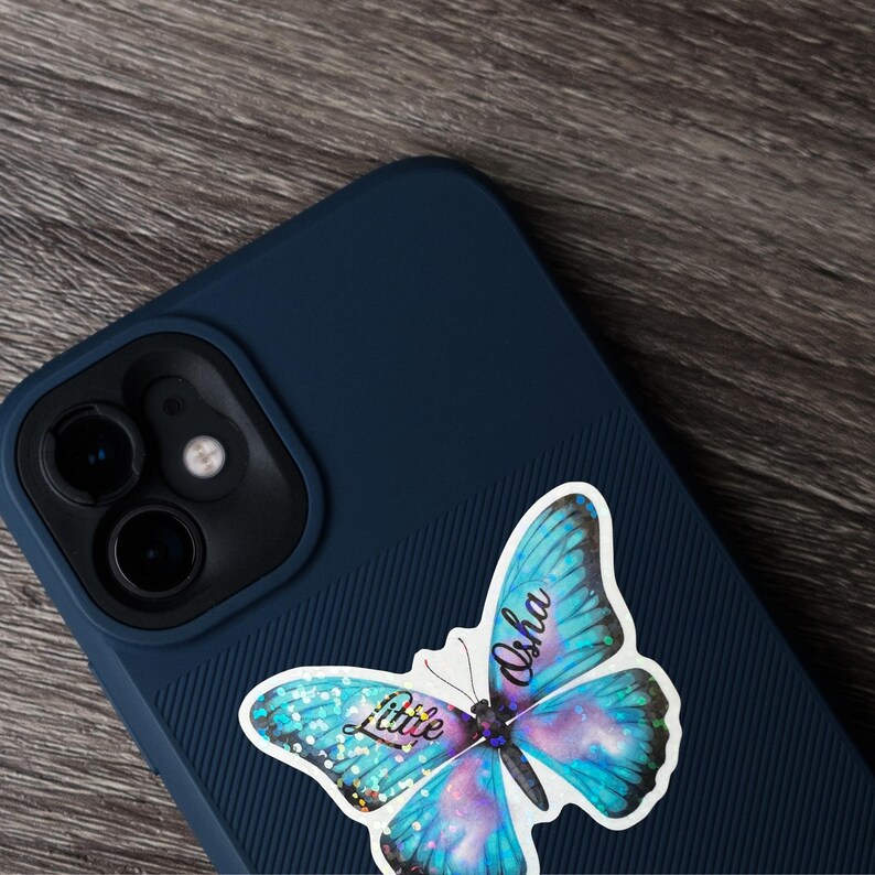May include: A blue phone case with a butterfly sticker. The sticker has a blue, purple, and white butterfly design with the words "Little Osha" in cursive. The phone case is dark blue and has a textured design.