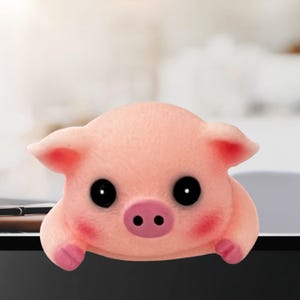 May include: A pink pig-shaped desk accessory with large black eyes, small pink ears, and rosy cheeks. The pig appears to be peeking over the edge of a desk or computer monitor. The overall design is cute and cartoonish.