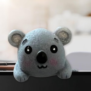 May include: A light gray koala figurine with a black nose, large black eyes, and pink cheeks. The koala is perched on a dark surface, with its paws visible. The figurine has a cute, cartoonish design.
