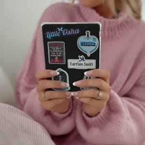 May include: A black tablet case adorned with colorful stickers. The stickers feature text like "Little Osha", "FISHER", and "Carrion Swift Logistics". The tablet is held by a person wearing a pink sweater.