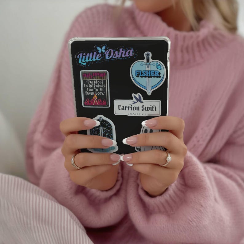 May include: A black tablet case adorned with colorful stickers. The stickers feature text like "Little Osha", "FISHER", and "Carrion Swift Logistics". The tablet is held by a person wearing a pink sweater.