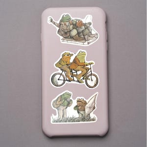 Puede incluir: Una funda de tel&eacute;fono rosa claro adornada con ilustraciones caprichosas de ranas. Las pegatinas representan ranas esquiando, montando en bicicleta t&aacute;ndem y de pie en un campo. Las ranas est&aacute;n vestidas con varios atuendos.