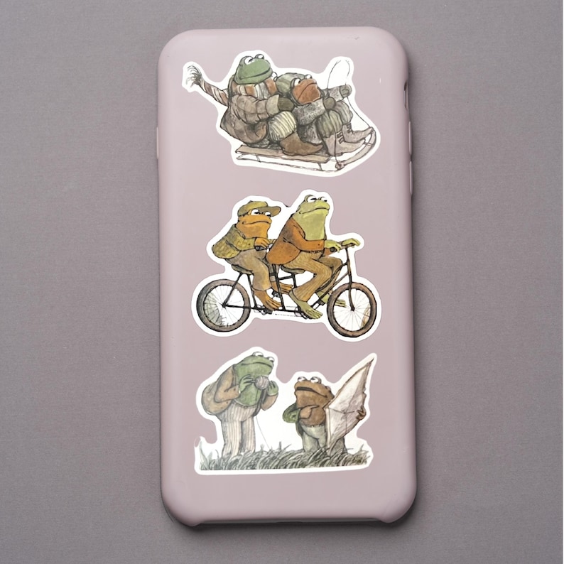 Puede incluir: Una funda de tel&eacute;fono rosa claro adornada con ilustraciones caprichosas de ranas. Las pegatinas representan ranas esquiando, montando en bicicleta t&aacute;ndem y de pie en un campo. Las ranas est&aacute;n vestidas con varios atuendos.