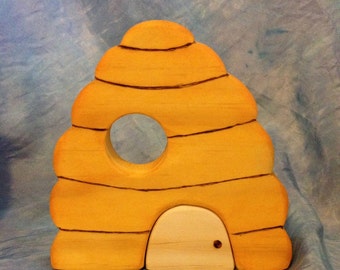 May include: A wooden beehive toy painted yellow with black stripes and a round opening. The bottom features a small, off-white door with a small brown dot. The toy is designed to resemble a real beehive.