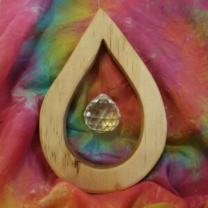 May include: A wooden teardrop-shaped frame with a clear crystal ball suspended within. The frame is crafted from light-coloured wood. The backdrop is a vibrant tie-dye fabric, featuring pink, orange, yellow, green, and blue shades.
