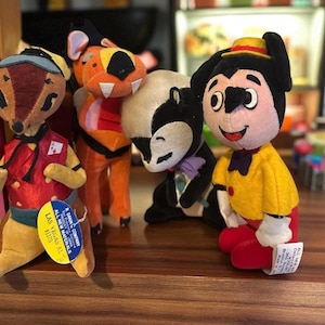 Pu&ograve; includere: Una collezione di peluche vintage, tra cui un cervo, una tigre, una puzzola e un personaggio con una camicia gialla e pantaloni rossi. Il cervo indossa un gilet rosso e un berretto blu. La tigre &egrave; arancione e rossa. La puzzola &egrave; bianca e nera.