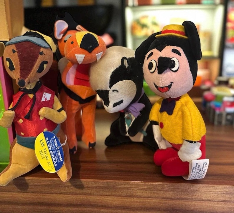 Pu&ograve; includere: Una collezione di peluche vintage, tra cui un cervo, una tigre, una puzzola e un personaggio con una camicia gialla e pantaloni rossi. Il cervo indossa un gilet rosso e un berretto blu. La tigre &egrave; arancione e rossa. La puzzola &egrave; bianca e nera.