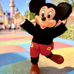 Puede incluir: Un peluche de Mickey Mouse, vestido con una camiseta negra, pantalones cortos rojos con botones amarillos y zapatos rojos grandes. El fondo presenta una pasarela colorida y un castillo, lo que sugiere un parque tem&aacute;tico.