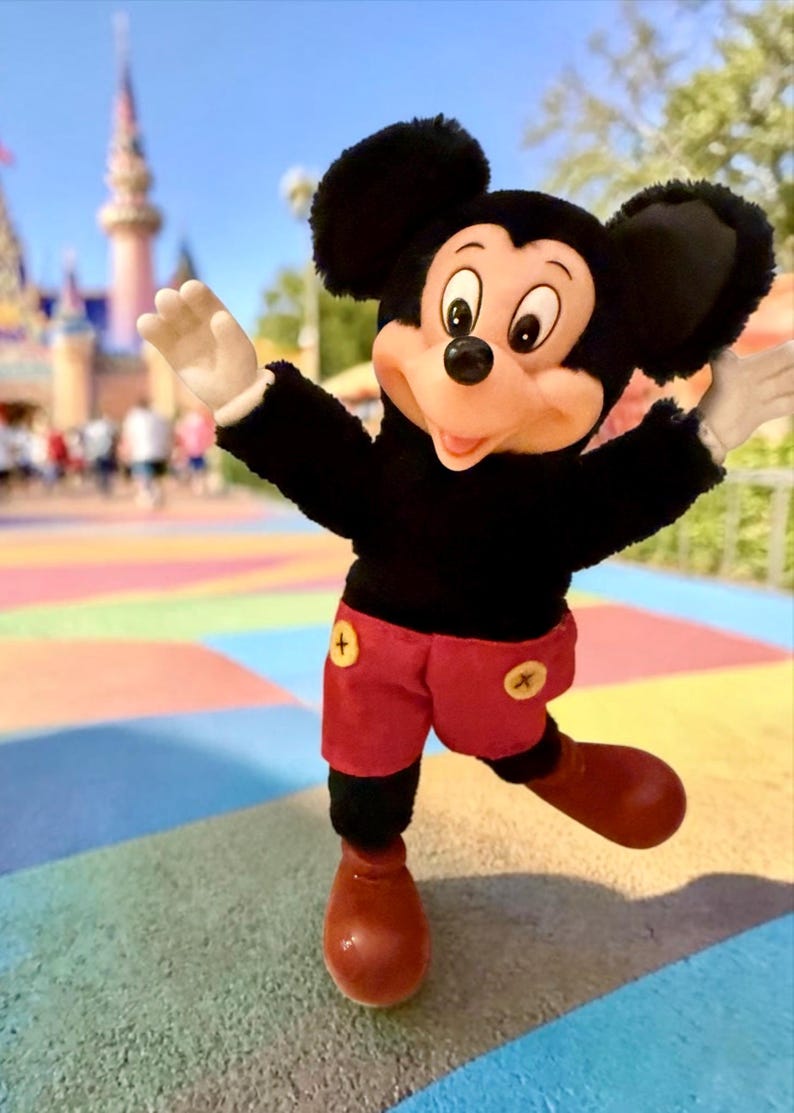 Puede incluir: Un peluche de Mickey Mouse, vestido con una camiseta negra, pantalones cortos rojos con botones amarillos y zapatos rojos grandes. El fondo presenta una pasarela colorida y un castillo, lo que sugiere un parque tem&aacute;tico.