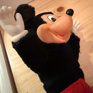 Puede incluir: Una marioneta de mano de Mickey Mouse con un cuerpo de felpa negro, una gran nariz naranja y guantes blancos. La marioneta tiene un atuendo rojo con un bot&oacute;n amarillo. Es un personaje cl&aacute;sico de Disney.