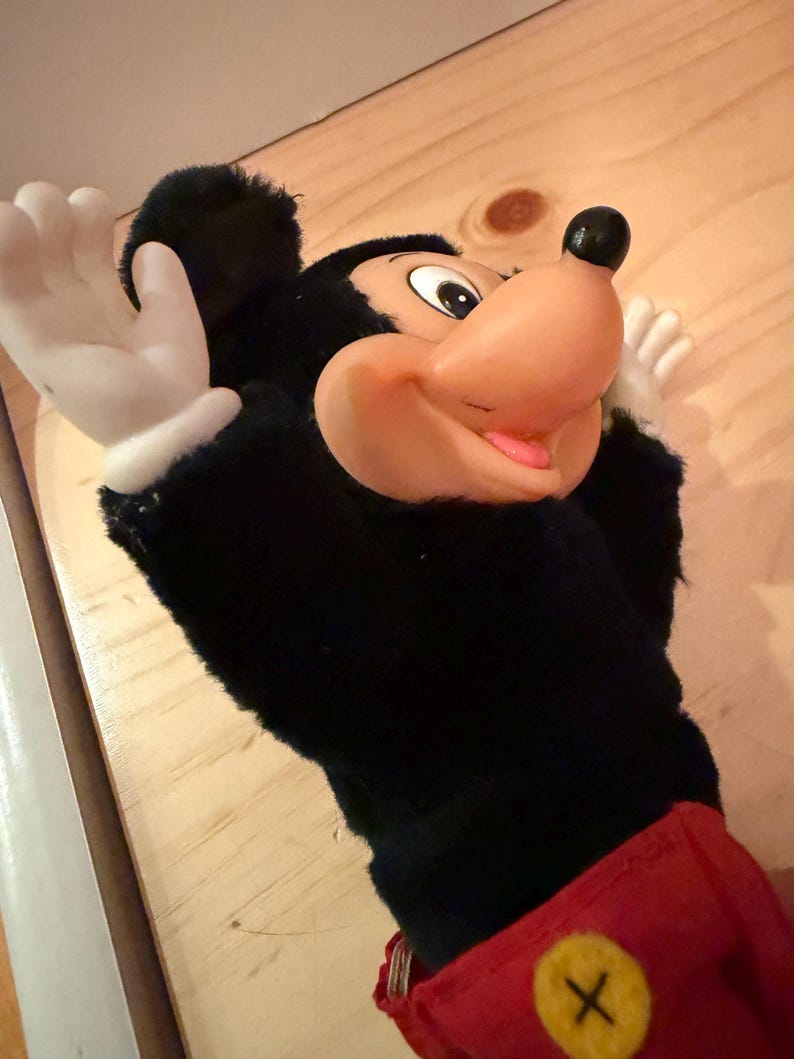 Puede incluir: Una marioneta de mano de Mickey Mouse con un cuerpo de felpa negro, una gran nariz naranja y guantes blancos. La marioneta tiene un atuendo rojo con un bot&oacute;n amarillo. Es un personaje cl&aacute;sico de Disney.