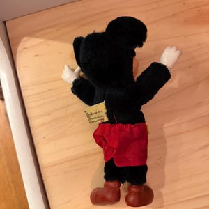 Puede incluir: Un peluche vintage de Mickey Mouse, visto desde atr&aacute;s. El peluche presenta un cuerpo negro, pantalones cortos rojos y zapatos marrones grandes. Los brazos y las manos son blancos. Una etiqueta est&aacute; adjunta. El juguete est&aacute; sobre una superficie de madera.