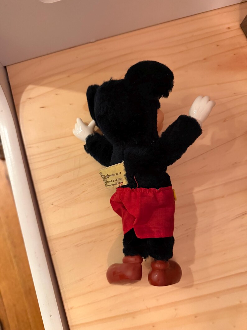 Puede incluir: Un peluche vintage de Mickey Mouse, visto desde atr&aacute;s. El peluche presenta un cuerpo negro, pantalones cortos rojos y zapatos marrones grandes. Los brazos y las manos son blancos. Una etiqueta est&aacute; adjunta. El juguete est&aacute; sobre una superficie de madera.