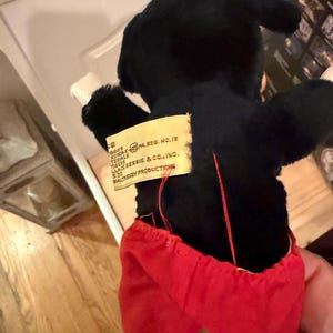 Puede incluir: Un juguete de peluche negro vintage con pantalones cortos rojos y guantes blancos. El juguete tiene una etiqueta que dice "KOREA PA.REG. NO.12" y "WALT DISNEY PRODUCTIONS". Los brazos del juguete est&aacute;n extendidos.