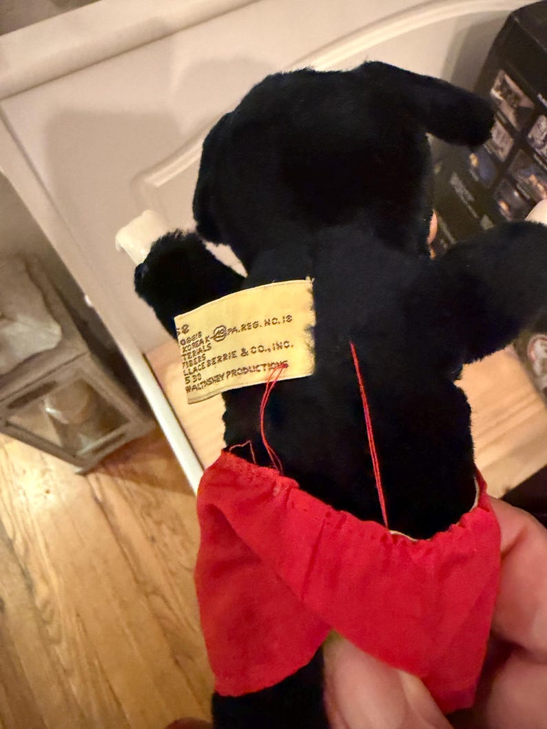 Puede incluir: Un juguete de peluche negro vintage con pantalones cortos rojos y guantes blancos. El juguete tiene una etiqueta que dice "KOREA PA.REG. NO.12" y "WALT DISNEY PRODUCTIONS". Los brazos del juguete est&aacute;n extendidos.
