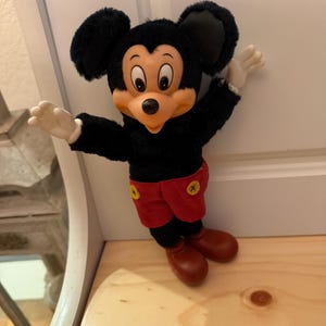 Puede incluir: Un peluche vintage de Mickey Mouse. El peluche presenta un cuerpo negro, grandes orejas negras, una cara bronceada, guantes blancos, pantalones cortos rojos con botones amarillos y zapatos marrones. El juguete est&aacute; posando con los brazos extendidos.