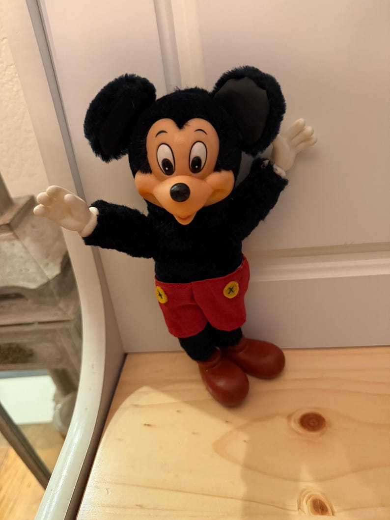 Puede incluir: Un peluche vintage de Mickey Mouse. El peluche presenta un cuerpo negro, grandes orejas negras, una cara bronceada, guantes blancos, pantalones cortos rojos con botones amarillos y zapatos marrones. El juguete est&aacute; posando con los brazos extendidos.
