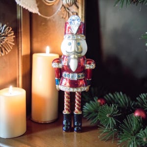 May include: A decorative Christmas nutcracker figurine. The figurine is red with gold and blue accents, and is adorned with rhinestones. It has a white face, a gold mustache, and black boots. The nutcracker is standing near lit candles and Christmas greenery.