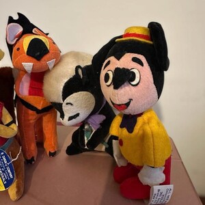 Pu&ograve; includere: Una collezione di peluche vintage, tra cui un personaggio dei cartoni animati con una camicia gialla, pantaloni rossi e un cappello. Altri peluche includono una tigre e una puzzola. I giocattoli sono fatti di tessuto morbido e hanno tratti del viso dettagliati.