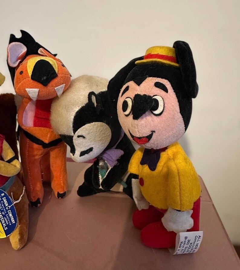 Pu&ograve; includere: Una collezione di peluche vintage, tra cui un personaggio dei cartoni animati con una camicia gialla, pantaloni rossi e un cappello. Altri peluche includono una tigre e una puzzola. I giocattoli sono fatti di tessuto morbido e hanno tratti del viso dettagliati.