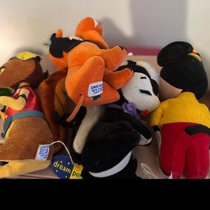 Pu&ograve; includere: Una collezione di peluche in vari colori. I giocattoli includono un personaggio marrone e giallo, un personaggio arancione e nero, un personaggio bianco e nero e un personaggio con una camicia gialla e pantaloni rossi. Alcuni giocattoli hanno etichette con la scritta "dream pets".