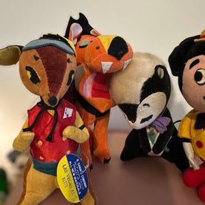 Pu&ograve; includere: Una collezione di giocattoli vintage di peluche a forma di animali. I giocattoli includono un cervo con un gilet rosso, un tasso in smoking e altri personaggi colorati. Il cervo ha un'etichetta che dice "R. Dakin Company, Las Vegas AL".