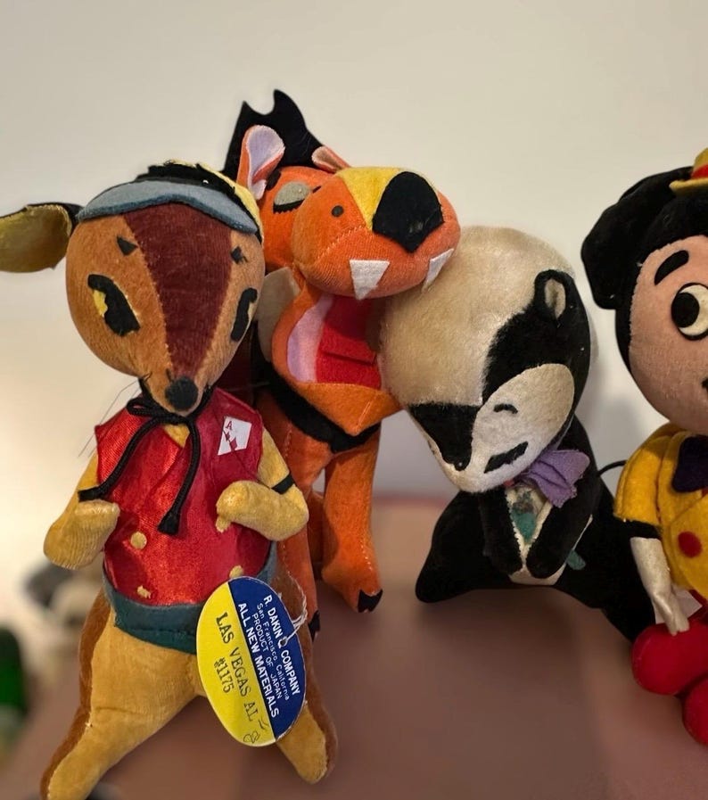Pu&ograve; includere: Una collezione di giocattoli vintage di peluche a forma di animali. I giocattoli includono un cervo con un gilet rosso, un tasso in smoking e altri personaggi colorati. Il cervo ha un'etichetta che dice "R. Dakin Company, Las Vegas AL".
