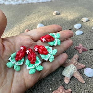 May include: Four handmade polymer clay earrings featuring a mermaid design. Each earring has a red hair, a white starfish, and a green and white base. The earrings are displayed on a sandy beach with seashells and starfish.