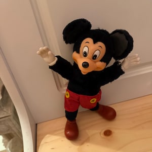 Puede incluir: Un peluche vintage de Mickey Mouse. El peluche tiene un cuerpo negro, orejas grandes negras, una cara beige y pantalones cortos rojos con botones amarillos. El juguete est&aacute; posando con los brazos extendidos y zapatos marrones.