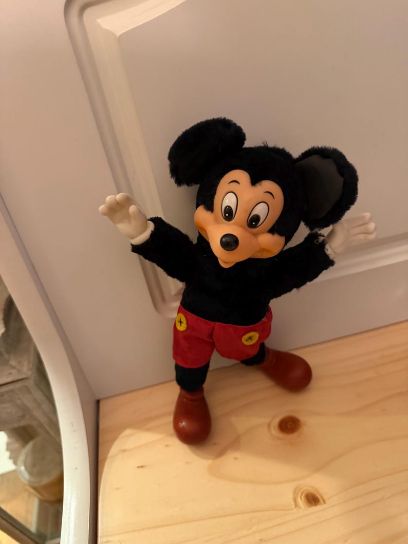 Puede incluir: Un peluche vintage de Mickey Mouse. El peluche tiene un cuerpo negro, orejas grandes negras, una cara beige y pantalones cortos rojos con botones amarillos. El juguete est&aacute; posando con los brazos extendidos y zapatos marrones.