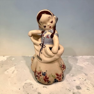 May include: A vintage ceramic figurine of a girl in a bonnet, holding a bag. The figurine is painted with a light color palette, featuring a blue dress and floral accents in red, blue, and green. The girl has closed eyes and brown hair.