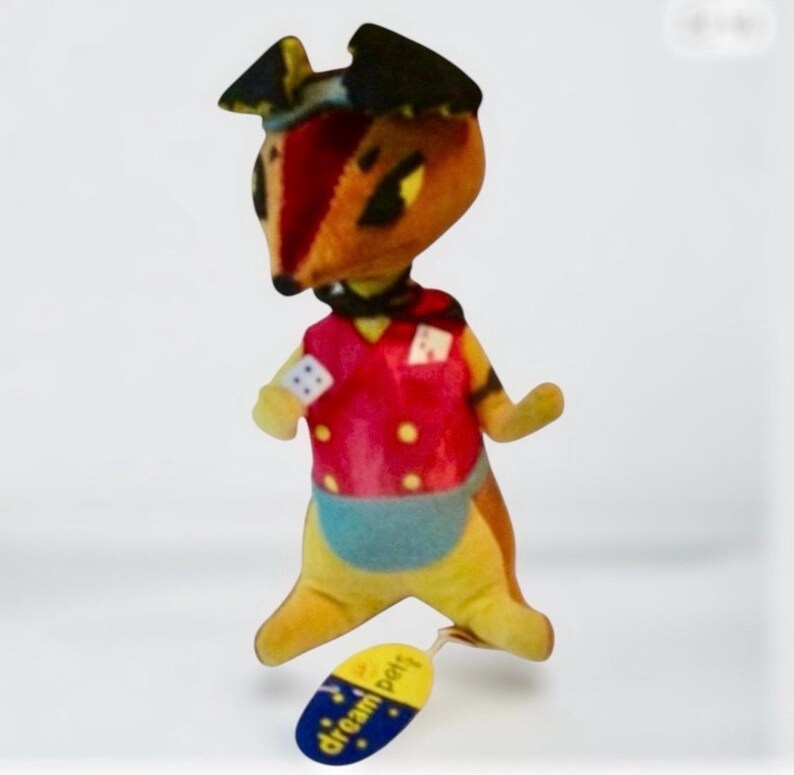 Pu&ograve; includere: Un peluche a forma di animale con un gilet rosso, pantaloncini blu e un cappello nero. Il giocattolo ha un'etichetta gialla con la scritta "dream pets". L'animale ha una faccia da volpe con occhi neri e naso rosso.