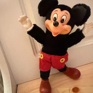 Puede incluir: Un peluche de Mickey Mouse con cuerpo negro, pantalones cortos rojos con botones amarillos y zapatos marrones grandes. El juguete tiene una cara sonriente, guantes blancos y orejas negras grandes. El juguete est&aacute; posando con los brazos extendidos.
