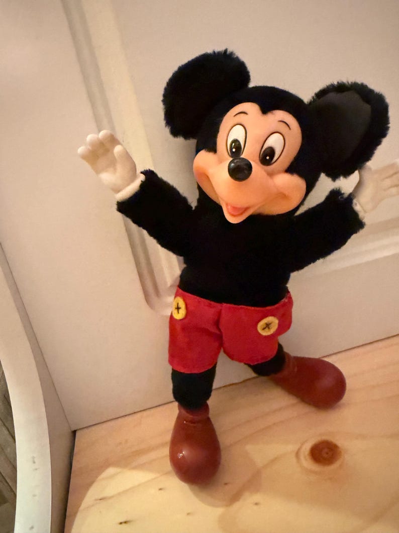 Puede incluir: Un peluche de Mickey Mouse con cuerpo negro, pantalones cortos rojos con botones amarillos y zapatos marrones grandes. El juguete tiene una cara sonriente, guantes blancos y orejas negras grandes. El juguete est&aacute; posando con los brazos extendidos.