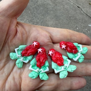 May include: Four handmade polymer clay mermaid figurines. Each figurine features a red-haired mermaid with a pink starfish hair accessory, a green tail, and a white top. The figurines are held in a person's hand.