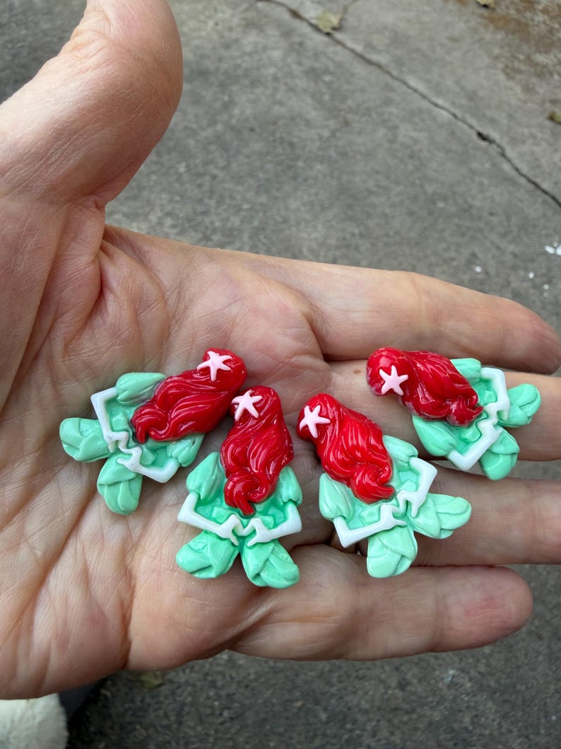 May include: Four handmade polymer clay mermaid figurines. Each figurine features a red-haired mermaid with a pink starfish hair accessory, a green tail, and a white top. The figurines are held in a person's hand.