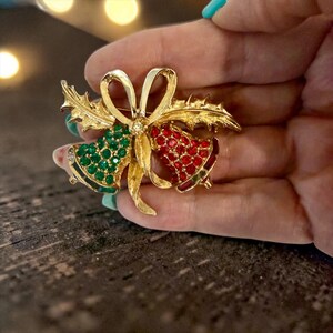 May include: A gold-tone brooch featuring two bells tied with a ribbon and holly leaves. One bell is encrusted with green rhinestones, while the other is covered in red rhinestones. The metal surface has a textured finish with a central clear stone.