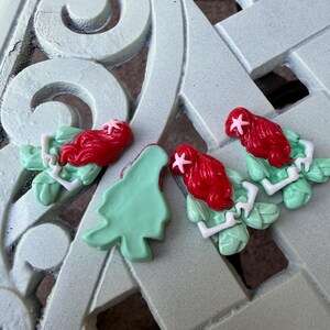 May include: Four resin mermaid-themed embellishments with red hair, green leaves, and white accents. One detached green leaf is also present. The items are arranged on a light green, patterned surface.