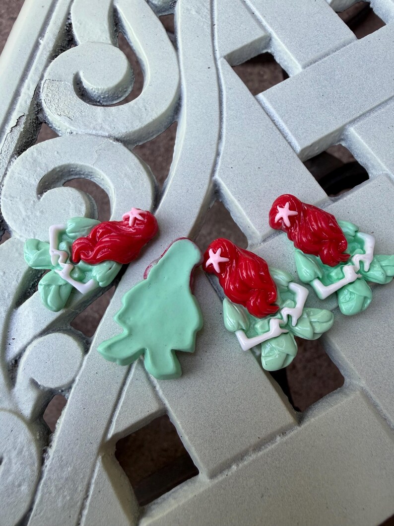 May include: Four resin mermaid-themed embellishments with red hair, green leaves, and white accents. One detached green leaf is also present. The items are arranged on a light green, patterned surface.