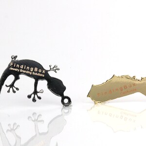 Puede incluir: Dos etiquetas met&aacute;licas con el texto "FindingBox Jewelry Branding Solutions" en letras doradas. Una etiqueta tiene forma de gecko y es negra, la otra tiene forma de estado y es dorada.