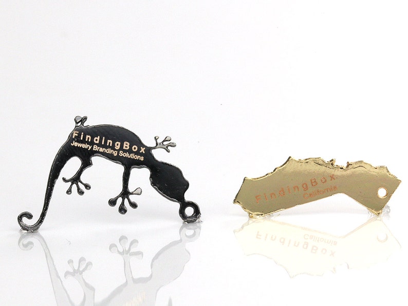 Puede incluir: Dos etiquetas met&aacute;licas con el texto "FindingBox Jewelry Branding Solutions" en letras doradas. Una etiqueta tiene forma de gecko y es negra, la otra tiene forma de estado y es dorada.