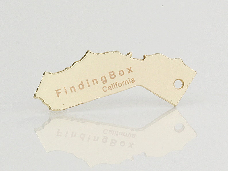 Puede incluir: Encanto de metal de color dorado con forma del estado de California con el texto "FindingBox California" en &eacute;l.