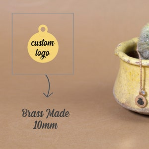 May include: A gold-colored brass tag with a round shape and a hole at the top. The tag is 10mm in diameter and has the text "custom logo" printed on it. The tag is shown next to a small potted cactus.