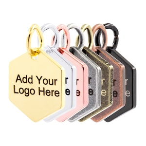 May include: A set of six hexagonal metal tags in different finishes: gold, rose gold, silver, gunmetal, antique brass, and black. Each tag has a hole at the top for attaching to a keychain or other item. The gold tag has the text "Add Your Logo Here" engraved on it.
