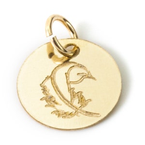 May include: Gold-toned metal charm with a circular shape and a loop at the top. The charm features an engraved design of a stylized feather.
