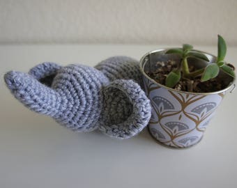 May include: A crocheted gray elephant toy laying on a white surface next to a small metal planter with a green succulent plant. The planter has a white and gold geometric pattern.