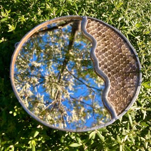 May include: A round mirror with a silver frame and a textured, honeycomb-like design on one side. The mirror reflects a blue sky and tree branches. The mirror is set against a backdrop of green foliage.