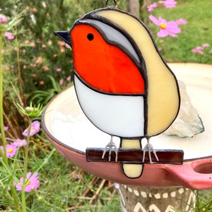 Può includere: Un ornamento di pettirosso in vetro colorato con petto rosso, pancia bianca e testa gialla. Il pettirosso è appollaiato su un ramo marrone.