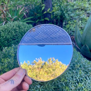 Può includere: Uno specchio rotondo portatile con una cornice in metallo argentato. La metà superiore dello specchio presenta un motivo strutturato a scaglie, mentre la metà inferiore riflette un cielo blu e un fogliame verde. Lo specchio è tenuto contro piante verdi.